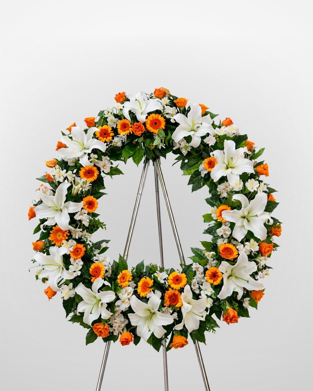 Aurora – Corona Fúnebre en Trípode de Lirios Blancos con Gerberas Naranjas y Mini Rosas, toque cálido y elegante