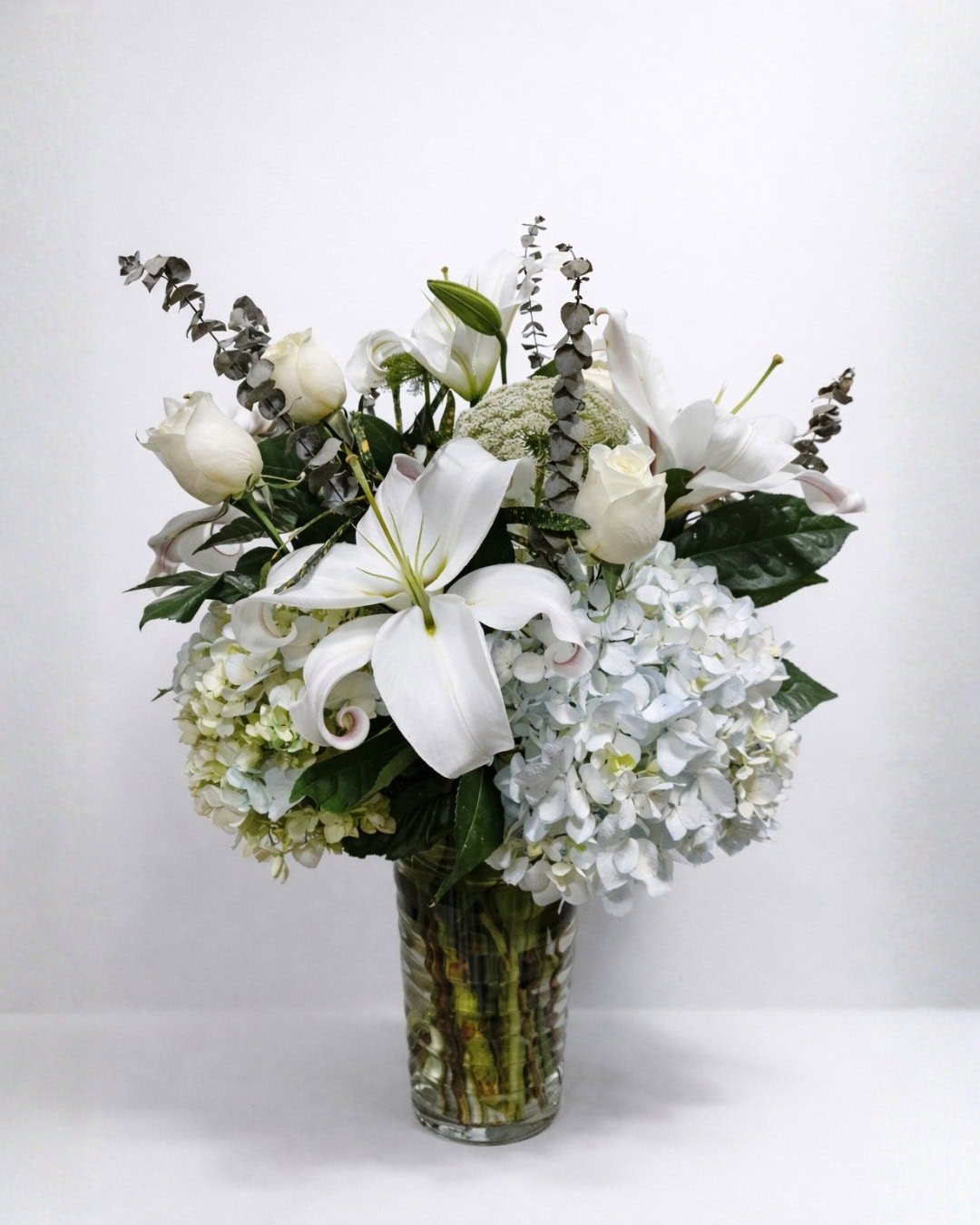 Nébula – Florero de vidrio de lirios blancos y rosas marfil con hortensias y eucalipto